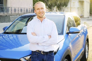 Genç adam onun yeni Karayip mavi araba yaslanmış beyaz gömlek gülümseyen portresi. Otomobil memnun yüz ifadesi, kentsel arka plan ile yakın duran yakışıklı sarışın iş adamı. Kopya alanı