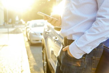 Smartphone kullanarak genç adam onun yeni Karayip mavi araba, eğilerek mavi gömlek resmi ware. Boş ekran cep telefonu elinde tutan bir erkek. GPS navigasyon kavramı. Yakın çekim, uzay, arka plan kopya.