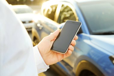 Akıllı telefon kullanan genç adam mavi gömlek resmi ware ayakta onun yeni Karayip mavi araba yakınındaki. Boş ekran cep telefonu elinde tutan bir erkek. GPS navigasyon kavramı. Yakın çekim, uzay, arka plan kopya.