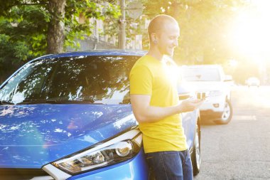 Sarı tişört yeni onun Karayip mavi araba eğilerek telefonla mesajlaşma genç adam. Yakışıklı erkek otomobil duran, smartphone kullanarak rahat kıyafetle uygun. Kopyalama alanı, arka plan, günbatımı