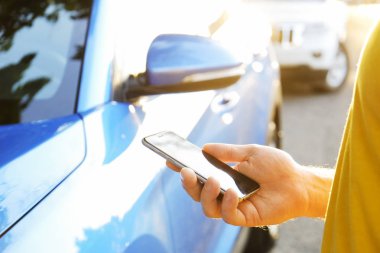 Smartphone kullanarak sarı tişört ayakta yakınındaki yeni Karayip mavi arabasını, Genç adam. Boş ekran cep telefonu elinde tutan bir erkek. GPS navigasyon kavramı. Yakın çekim, kopya alanı, arka plan, günbatımı