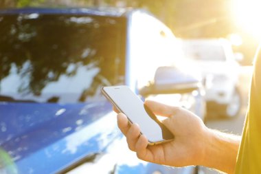 Smartphone kullanarak sarı tişört ayakta yakınındaki yeni Karayip mavi arabasını, Genç adam. Boş ekran cep telefonu elinde tutan bir erkek. GPS navigasyon kavramı. Yakın çekim, kopya alanı, arka plan, günbatımı