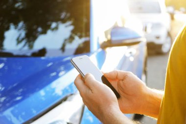 Smartphone kullanarak sarı tişört ayakta yakınındaki yeni Karayip mavi arabasını, Genç adam. Boş ekran cep telefonu elinde tutan bir erkek. GPS navigasyon kavramı. Yakın çekim, kopya alanı, arka plan, günbatımı