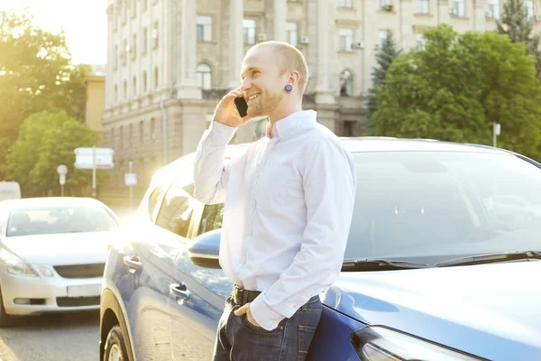 Smartphone kullanarak genç adam onun yeni Karayip mavi araba, eğilerek mavi gömlek resmi ware. Otomobil yakınındaki telefonda konuşurken iş adamı. GPS navigasyon kavramı. Yakın çekim, uzay, arka plan kopya.