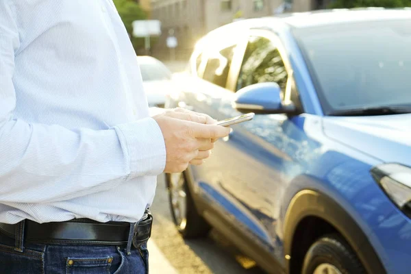 Akıllı telefon kullanan genç adam mavi gömlek resmi ware ayakta onun yeni Karayip mavi araba yakınındaki. Boş ekran cep telefonu elinde tutan bir erkek. GPS navigasyon kavramı. Yakın çekim, uzay, arka plan kopya.