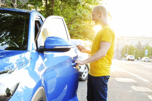 Smartphone kullanarak sarı tişört açılış kapı yeni Karayip mavi arabanın, Genç adam. Ellerini arabaya, Erkek holding cep telefonu. GPS navigasyon kavramı. Kopyalama alanı, arka plan, günbatımı.