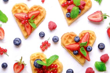 Çeşitli çilek karışımı, çilek, böğürtlen, ahududu ile kalp şekli Belçika Waffle ve nane yaprağı beyaz seramik plaka üzerinde kırmızı kuş üzümü süslenmiş. Yakın çekim, kopya alanı, arka plan, üstten görünüm.