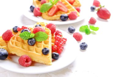 Çeşitli çilek karışımı, çilek, böğürtlen, ahududu ile kalp şekli Belçika Waffle ve nane yaprağı beyaz seramik plaka üzerinde kırmızı kuş üzümü süslenmiş. Yakın çekim, kopya alanı, arka plan, üstten görünüm.