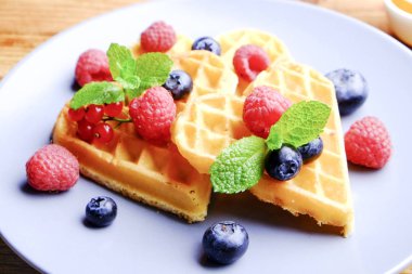 Çeşitli çilek karışımı, çilek, böğürtlen, ahududu ile kalp şekli Belçika Waffle ve nane yaprağı beyaz seramik plaka üzerinde kırmızı kuş üzümü süslenmiş. Yakın çekim, kopya alanı, arka plan, üstten görünüm.