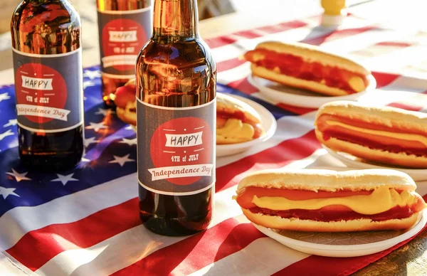 Fourth of July table setting, beer bottle with original label design, Happy Independence day, classic hot dog with catchup and mustard, American flag. Outdoors recreation area. Close up, background.