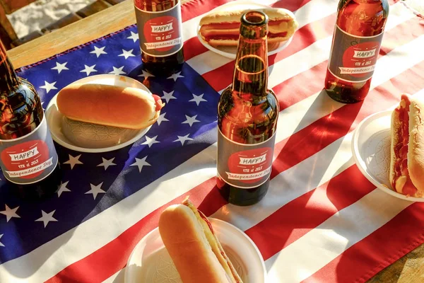 Fourth of July table setting, beer bottle with original label design, Happy Independence day, classic hot dog with catchup and mustard, American flag. Outdoors recreation area. Close up, background.