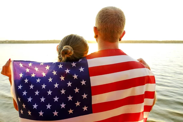 Rear view of young American patriotic couple hugging with USA flag on their backs enjoying beautiful summer sunset by the river. Independence day celebration concept. Background, close up, copy space.
