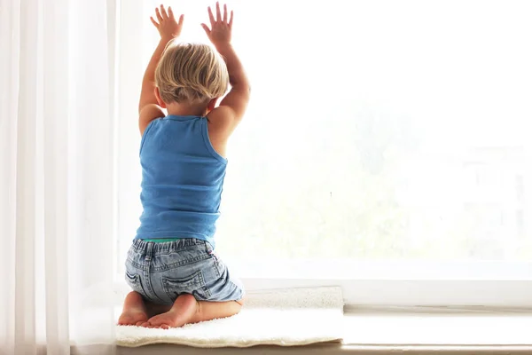 Child missing his mother concept. Little cute blond three year old boy sitting on windowsill touching window glass, looking sad. Kid in blue tank top playing alone. Background, close up, copy space.
