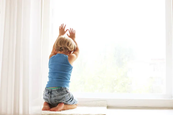 Child missing his mother concept. Little cute blond three year old boy sitting on windowsill touching window glass, looking sad. Kid in blue tank top playing alone. Background, close up, copy space.