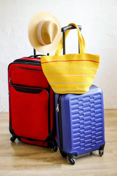 Red textile suitcase & blue hard shell luggage, extended telescopic handle, straw hat, yellow beach bag, white decorative texture wall background. Couples retreat trip concept. Close up, copy space.