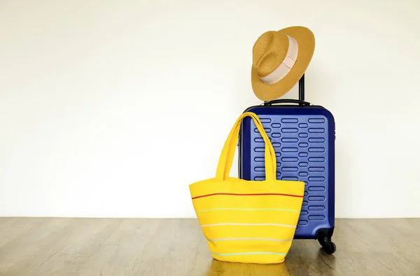 Single blue plastic hard shell luggage with women\'s straw hat hanging on extended telescopic handle and yellow beach bag. One suitcase prepared for vacation trip. White wall background, copy space.