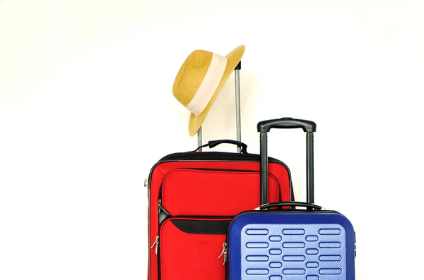 Red textile suitcase & blue hard shell luggage with straw hat hanging on extended telescopic handle, white decorative texture wall background. Couples retreat trip concept. Close up, copy space.