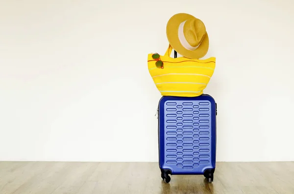 Single blue plastic hard shell luggage with women\'s straw hat hanging on extended telescopic handle, yellow beach bag & mirrored sunglasses. Suitcase for one concept. White wall background, copy space