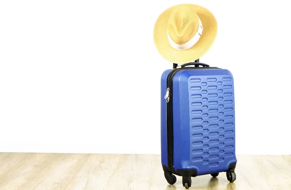 Single blue plastic hard shell luggage with woman straw hat hanging on extended telescopic handle. One suitcase. Traveling alone concept. White textured wall background, copy space for text, close up.