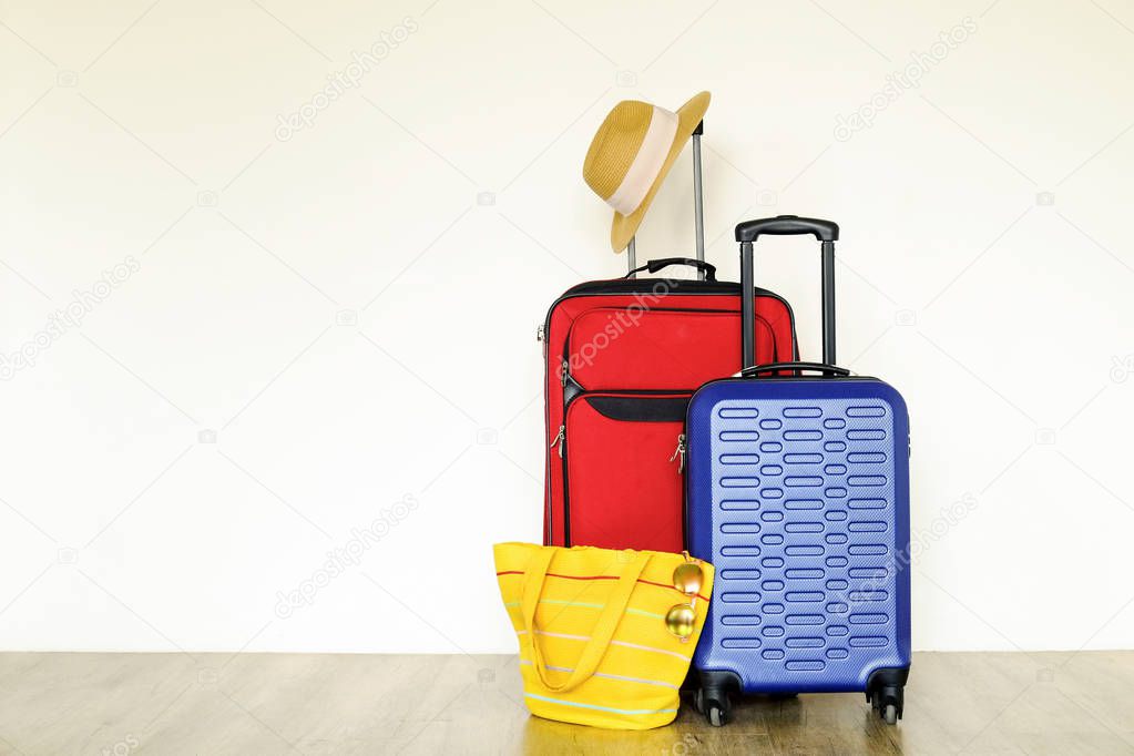 Red textile suitcase & blue hard shell luggage, extended telescopic handle, straw hat, yellow beach bag, mirrored sunglasses, white wall background. Couples retreat trip concept. Close up, copy space.