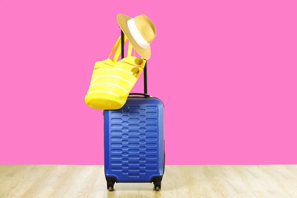 Single blue plastic hard shell luggage with women\'s straw hat hanging on extended telescopic handle, yellow beach bag, sunglasses. One suitcase prepared for vacation trip. Pink background, copy space