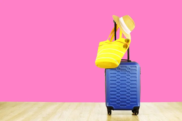Single blue plastic hard shell luggage with women's straw hat hanging on extended telescopic handle, yellow beach bag, sunglasses. One suitcase prepared for vacation trip. Pink background, copy space