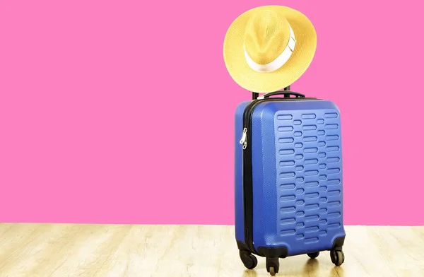 Single blue plastic hard shell luggage with woman straw hat hanging on extended telescopic handle. One suitcase. Traveling alone concept. Bright pink wall background, copy space for text, close up.