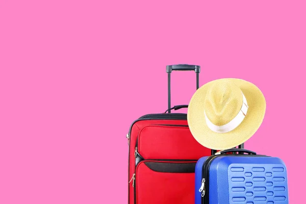 Red textile suitcase & blue hard shell luggage with straw hat hanging on extended telescopic handle, bright solid pink wall background. Couples retreat trip concept. Close up, copy space.