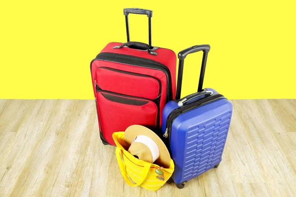 Red textile suitcase & blue hard shell luggage, extended telescopic handle, straw hat, yellow beach bag, mirrored sunglasses, yellow wall background. Couples retreat trip concept. Close up, copy space