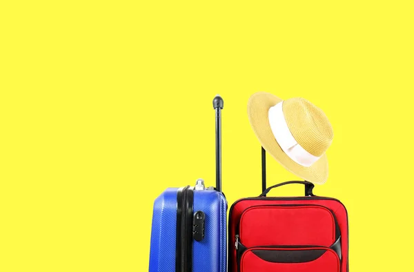 Red textile suitcase & blue hard shell luggage with straw hat hanging on extended telescopic handle, bright yellow isolated background. Couples retreat trip concept. Close up, copy space.