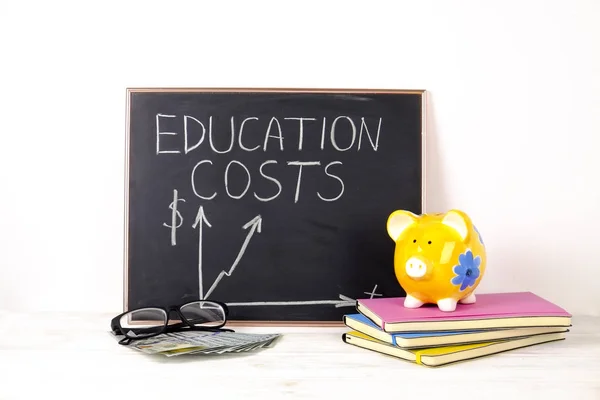 Growing education costs concept. Yellow piggy bank standing on colorful notebooks and stack of new crispy one hundred dollar bills. School supplies, blank blackboard, clock. Background, copy space.