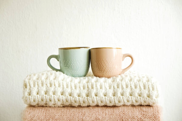 Two cups of coffee standing on bunch of knitted warm pastel color sweaters w/ different knitting patterns folded in stack. Fall winter knitwear clothing. Textured wall background. Close up, copy space