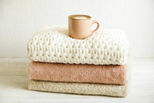 Cup of cappuccino coffee on bunch of knitted warm pastel color sweaters w/ different knitting patterns folded in stack. Fall winter knitwear clothing. Textured wall background. Close up, copy space.