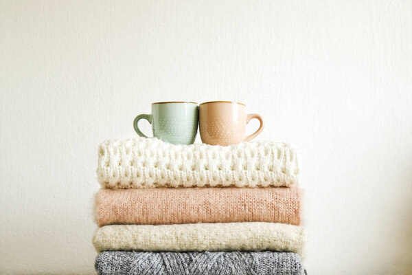 Two cups of coffee standing on bunch of knitted warm pastel color sweaters w/ different knitting patterns folded in stack. Fall winter knitwear clothing. Textured wall background. Close up, copy space
