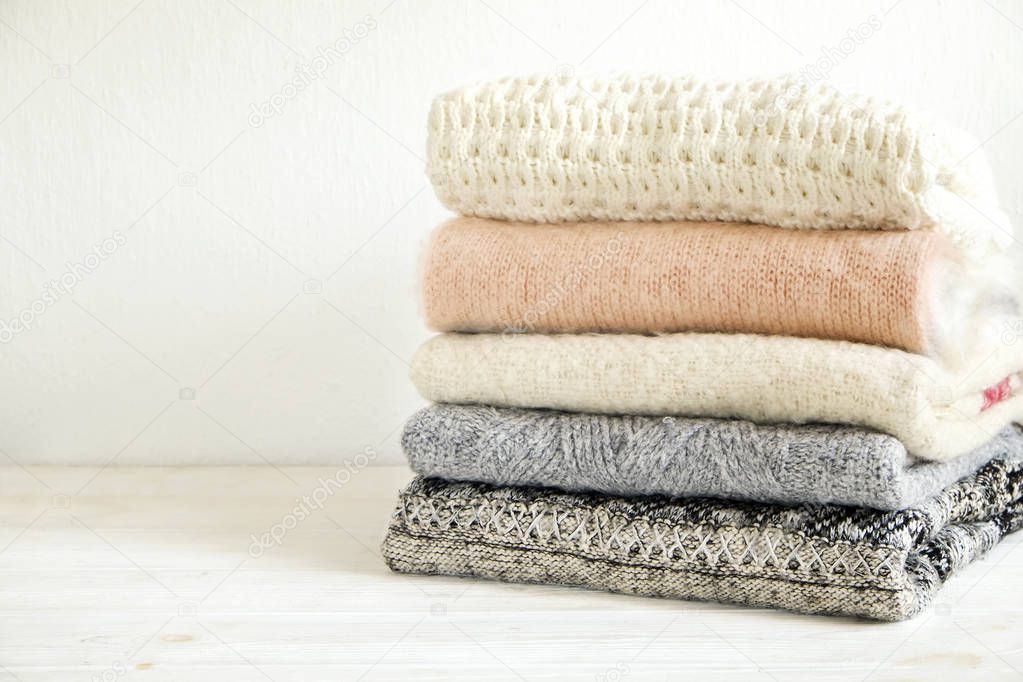 Bunch of knitted warm pastel color sweaters with different knitting patterns folded in stack on white wooden table, textured wall background. Fall winter season knitwear. Close up, copy space for text