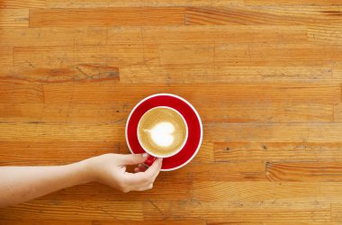 Kırmızı kalp şekli köpük latte sanat ile kahve fincan holding genç kadın eller en iyi görünümü. Kadın kapuçino içiyor, oturma grunged tarafından ahşap bar tablo çizmiş. Arka plan, yakın çekim, alanı Kopyala.
