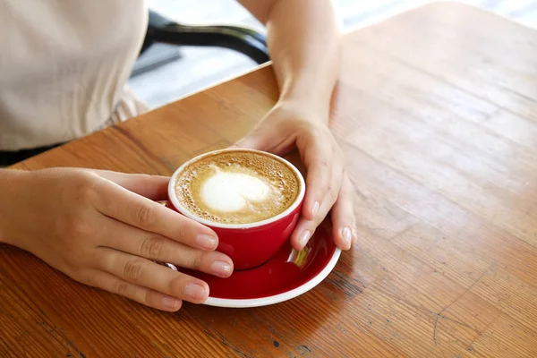 Kırmızı kalp şekli köpük latte sanat ile kahve fincan holding genç kadın eller en iyi görünümü. Kadın kapuçino içiyor, oturma grunged tarafından ahşap bar tablo çizmiş. Arka plan, yakın çekim, alanı Kopyala.