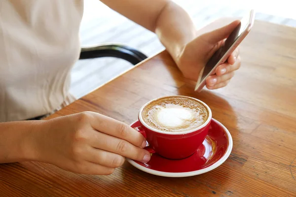 Young woman sitting at coffee place, having red cup of cappuccino with heart shape foam latte art. Female holding blank screen smartphone over wooden table. Background, close up, copy space, top view