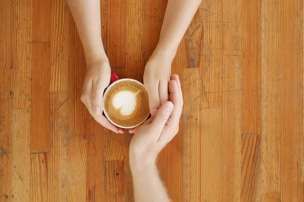 Young couple in love holding hands over red cup of coffee with heart shaped foam latte art on vintage grunged scratched table. Man & woman body language. Background, top view, close up, copy space.