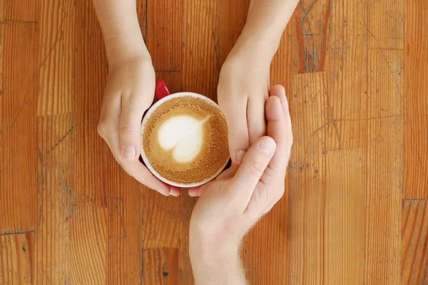 Young couple in love holding hands over red cup of coffee with heart shaped foam latte art on vintage grunged scratched table. Man & woman body language. Background, top view, close up, copy space.