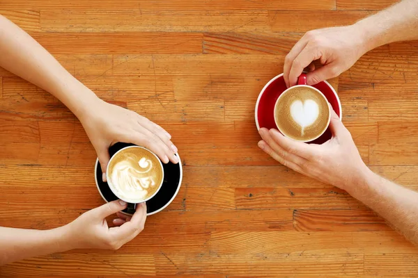 Young couple on a date, having cappuccino in vintage black and red cups heart and swan latte art, sitting at grunged scratched wooden table of coffee bar. Background, close up, top view, copy space.