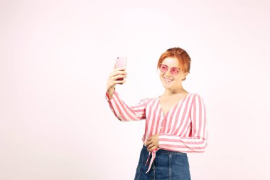 Self obsessed funny young beautiful woman with natural red hair, wearing pink cat eye sunglasses & stripped knot shirt taking selfie on cell phone. Wide sincere smile. Background, copy space, close up clipart