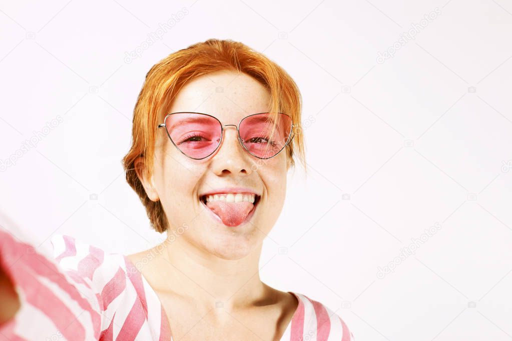 First person view selfie shot of funny young beautiful woman showing tongue, natural red hair, wearing pink cat eye sunglasses and stripped shirt. Wide sincere smile. Background, copy space, close up.