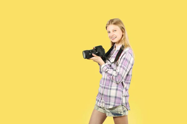 Young teenage girl holding digital photo camera with big lens & strap, taking pictures, smiling. Beautiful blond female photographer in checkered plaid shirt posing w/ dslr gear. Copy space background