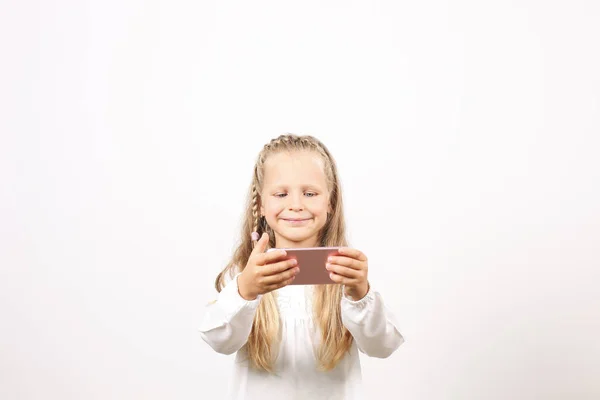 Portrait of positive & funny four year old blonde little girl, holding mobile cell phone, smiling at camera, taking selfies. Childhood & technology concept. Isolated background, copy space, close up.