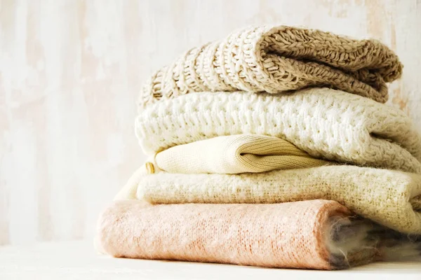 Bunch of knitted warm pastel color sweaters, different knitting patterns folded in messy stack on brown wooden table, grunged texture wall background. Fall winter season knitwear. Close up, copy space