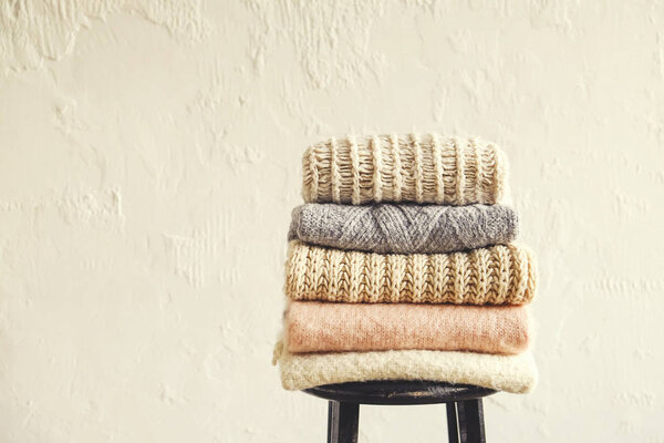 Bunch of knitted pastel color sweaters, different knitting patterns folded in neat stack on black wooden barstool, grunged texture wall background. Fall winter season knitwear. Close up, copy space.