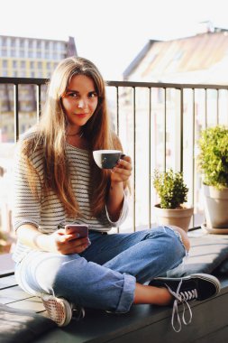 Cep telefonu üzerinde çatı üst teras kafe, hipster kafede sıcak cappuccino içecek içme tutan lotus poz, katlanmış bacaklar ile oturan güzel genç kadın. Arka plan, yakın çekim, alanı Kopyala.