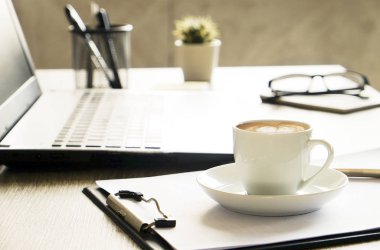 Boş ekran dizüstü bilgisayar, fincan cappuccino kahve, kaktüs, sarf malzemeleri ve katlanmış gözüne gözlük güneş ışığı tam geniş ofiste ahşap masa üzerinde. Yaratıcı çalışma alanı. Yakın çekim, uzay, arka plan kopya
