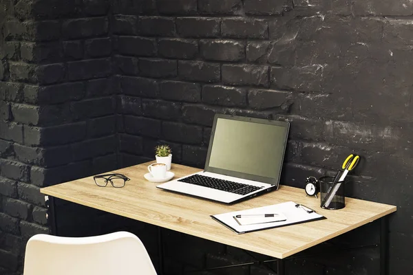 Creative workspace of a blogger. White laptop computer & folded eye glasses on wooden table in loft style office with black brick walls. Designer\'s table concept. Close up, copy space, background.